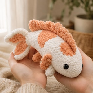 May include: A plush fish toy crafted from soft, white and peach-colored yarn. The fish features a textured, crocheted surface, a prominent dorsal fin, a bifurcated tail, and small side fins. It has two round, black plastic eyes on its head.