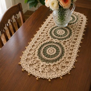 May include: An oval crochet table runner featuring cream-colored yarn with intricate lace patterns and three circular motifs in sage green. The edges are adorned with small, translucent amber-colored beads, creating a decorative border.