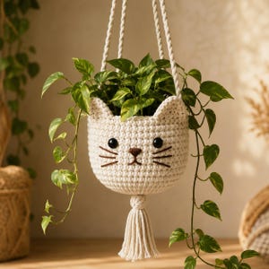 May include: A cream-coloured crocheted hanging planter shaped like a cat face with black button eyes, a brown embroidered nose, and whiskers. The planter features a matching cream-coloured tassel at the bottom and is suspended by three thick ropes.