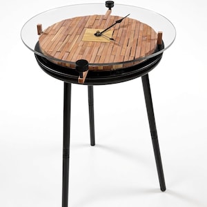 May include: A round table with three black legs and a black frame. The tabletop features a circular clock face made of small, light brown wooden slats arranged in a pattern, covered by a clear glass top with black clock hands and black accents.