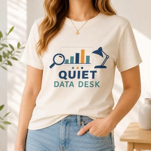 May include: A cream-coloured short-sleeve t-shirt featuring a graphic print of a magnifying glass, a bar chart, and a desk lamp. Below the graphics, the text reads "QUIET DATA DESK" in dark blue and teal lettering, centred on the chest.