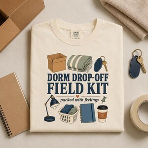 May include: A cream-colored t-shirt featuring a graphic print with the text DORM DROP-OFF FIELD KIT and packed with feelings. The illustration includes a cardboard box, a striped blanket, a key, a desk lamp, a laundry basket, a notebook, and a coffee cup.