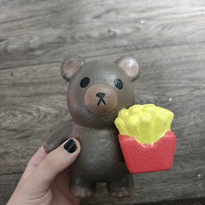 May include: A brown bear figurine with black eyes and a black nose, holding a red container filled with yellow french fries. The bear has a smooth, painted texture and rounded ears, standing in a vertical position with its arms extended forward.