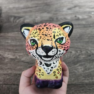 May include: A small, hand-painted figurine of a cheetah cub with a rounded head and large ears. The figure features a vibrant gradient of yellow, orange, and pink fur with black spots, bright green eyes, and a dark purple base at the bottom.