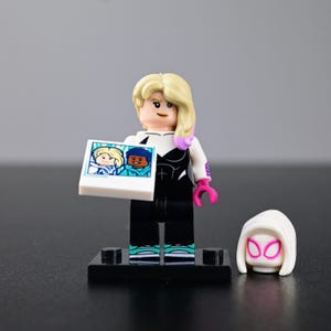 Op de afbeelding: Een LEGO-minifiguur met blond haar en roze highlights, gekleed in een zwart-wit pak, die een vierkant tegeltje vasthoudt met een cartoonafbeelding van twee figuren. Een losse witte capuchon met roze omlijnde oogopeningen ligt rechts.