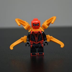 Peut inclure: Une figurine LEGO Spider-Man avec une tête rouge à motifs de toile noire, un torse noir et rouge avec un emblème d'araignée, et quatre bras mécaniques orange s'étendant du dos. La figurine a des bras et des jambes noirs avec des accents rouges.
