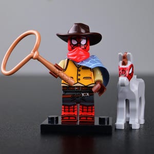 Peut inclure: Une figurine de style Lego habillée en cow-boy avec un chapeau marron, un bandana rouge, un gilet jaune et une cape bleue à motifs, tenant un lasso beige. Le personnage porte un masque rouge à motifs de toile d'araignée noire près d'un cheval blanc.