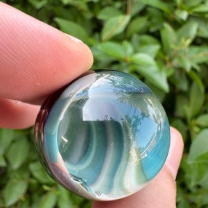 May include: A smooth, spherical glass marble featuring swirling bands of translucent teal, deep blue, and opaque white colors. The surface is highly reflective, showing light glints and distorted reflections of the surrounding environment.