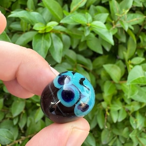 May include: A small, rounded glass bead featuring a deep black base color with a prominent, layered eye-like design. The pattern consists of a dark blue center surrounded by a bright turquoise ring, creating a distinct circular motif on the surface.