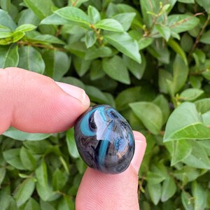 May include: A small, smooth, rounded glass bead featuring a dark, translucent base with swirling patterns of vibrant turquoise blue and deep black. The object has a polished, reflective surface that catches light, creating a glossy appearance.