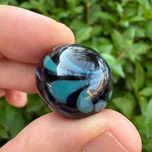 May include: A spherical glass marble featuring a deep black base color with swirling patterns of translucent teal and light blue glass. The surface is smooth and reflective, showing light highlights across the rounded, polished glass orb.