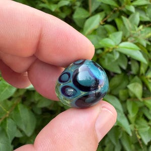 May include: A small, rounded glass bead featuring a translucent teal and light blue base with dark navy blue circular patterns embedded within the surface. The glass has a smooth, polished finish that reflects light across its curved, organic shape.