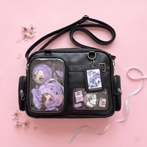 Peut inclure: Un sac ita noir avec une bandoulière réglable, doté de deux fenêtres de présentation transparentes à l'avant. La fenêtre gauche contient deux poupées en peluche violettes, tandis que la droite contient des cartes et un porte-badge.