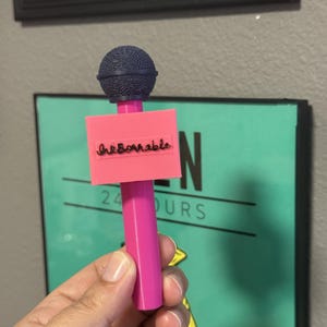 Puede incluir: Un accesorio de micrófono de plástico rosa con una cabeza esférica texturizada de color azul oscuro y una bandera rectangular rosa unida al mango. La bandera muestra la palabra "InkBornable" en letras cursivas negras en su parte frontal.