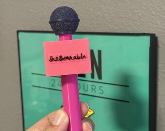 Può includere: Un accessorio a forma di microfono in plastica rosa con una testa sferica testurizzata blu scuro e una bandierina rettangolare rosa attaccata al manico. La bandierina mostra la parola "InkBornable" in corsivo nero sulla parte anteriore.