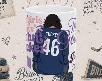 Könnte beinhalten: Ein weißer Keramikbecher mit einer Grafik einer Person mit dunklem, lockigem Haar von hinten, die ein dunkelblaues Eishockeytrikot mit dem Namen TUCKER und der Nummer 46 in Weiß trägt. Der Text lautet John Tucker, girls love und The Goal.