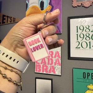 May include: A rectangular keychain shaped like a small book with a light pink border and a white center. The text BOOK LOVER is embossed in pink capital letters on the front. The keychain is attached to a silver metal split ring by a short chain.