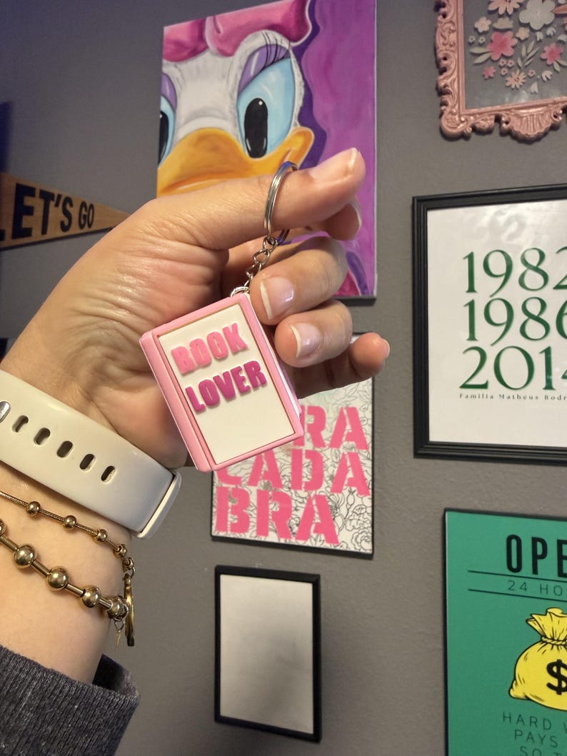May include: A rectangular keychain shaped like a small book with a light pink border and a white center. The text BOOK LOVER is embossed in pink capital letters on the front. The keychain is attached to a silver metal split ring by a short chain.