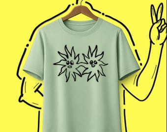 Könnte beinhalten: Ein helles, salbeigrünes Kurzarm-T-Shirt mit einer schwarzen Strichgrafik, die zwei miteinander verbundene sonnenartige Figuren mit einfachen Gesichtern zeigt. Das T-Shirt hängt an einem Holzkleiderbügel vor einem gelben Hintergrund.