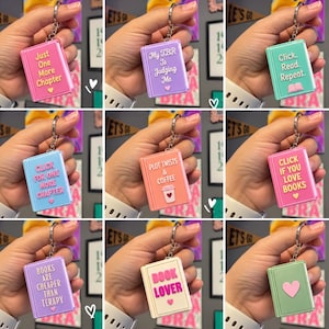 May include: A collection of nine rectangular book-shaped keychains in various pastel colors, each featuring a metal ring and unique text or symbols including phrases like Just One More Chapter, My TBR Is Judging Me, and Book Lover with heart icons.