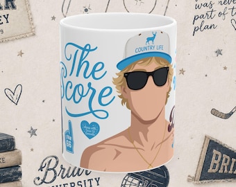 Könnte beinhalten: Ein weißer Keramikbecher mit einer grafischen Illustration einer Person mit blondem Haar, die eine schwarze Sonnenbrille und eine graue Kappe mit einem blauen Hirschlogo und dem Text "COUNTRY LIFE" trägt. Der Becher zeigt den blauen Text "The Score".