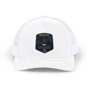Op de afbeelding: Een witte truckerpet met een donkerblauw schildvormig embleem op de voorzijde. Het embleem toont een kaart van de Madison River in Montana, inclusief de tekst Madison River, Montana, coördinaten, een visicoon en een afbeelding van een vismolen.