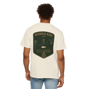 May include: An off-white short-sleeve t-shirt featuring a dark green shield-shaped graphic on the back. The graphic displays a map of the Deschutes River in Oregon, including text labels for Warm Springs, Trout Creek, and Sherars Falls.