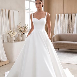 May include: A floor-length white wedding dress featuring a fitted bodice with a sweetheart neckline, thick shoulder straps, and a full, voluminous A-line skirt with integrated side pockets and a long train that extends across the floor.