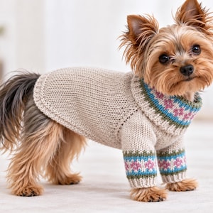 Puede incluir: Un perro pequeño con un suéter de punto beige de cuello alto. El suéter presenta una banda decorativa con patrones geométricos en azul, verde y rosa alrededor del cuello y los puños de las mangas delanteras, con textura de punto acanalado.