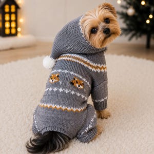 May include: A gray knit dog sweater with a hood and a white pom-pom. The sweater features a repeating pattern of small orange and white fox faces, along with horizontal white and mustard yellow stripes across the back and sleeves.