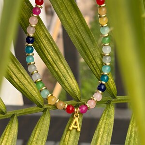 May include: A beaded bracelet featuring a variety of multicolored round beads in shades of pink, red, yellow, green, blue, and white, interspersed with small gold-toned metal spacers. A gold-toned metal charm in the shape of the letter A hangs centrally.