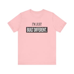 Könnte beinhalten: Ein hellrosa kurzärmeliges T-Shirt mit dem Text I'M JUST BUILT DIFFERENT. Die Wörter I'M JUST sind in Schwarz gedruckt, während die Wörter BUILT DIFFERENT. in Weiß innerhalb eines soliden schwarzen Rechtecks auf der Brust zu sehen sind.