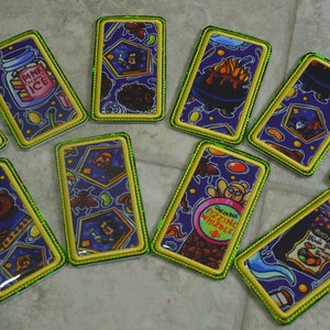 May include: A collection of rectangular patches with various colorful designs. Each patch has a purple background with cartoon illustrations and text, framed by a yellow border with green stitching. The patches feature images of potions, food, and creatures.