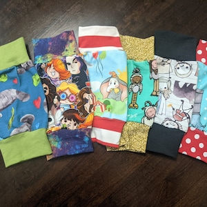 May include: Six colorful fabric leg warmers with different cartoon character designs. The leg warmers are made of a soft, stretchy fabric and have a ribbed cuff.