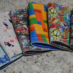 May include: Six different colorful zippered wallets with various patterns, including a blue and white Stitch pattern, a white and pink dog pattern, a black and white floral pattern, a colorful Lego pattern, a colorful cartoon pattern, and a black and white stained glass pattern.