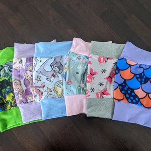 Op de afbeelding: Zes paar kleurrijke stoffen shorts met verschillende patronen, waaronder een jungle scène, een prinses, een dinosaurus, bloemen, ninjas en een zeemeermin. De shorts zijn allemaal gemaakt van een zachte, rekbare stof en hebben een comfortabele elastische tailleband.