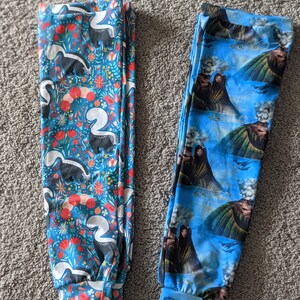 May include: Two pairs of blue leggings with different patterns. One pair features a white and black skunk pattern on a blue background with red and pink flowers. The other pair features a blue and green mountain scene with a volcano and people.