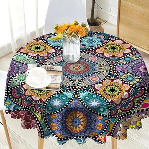 May include: A round tablecloth featuring a vibrant, multi-colored mandala pattern with intricate floral and geometric shapes in shades of orange, blue, purple, and green. A white ceramic cup and a small book rest on the center of the table.