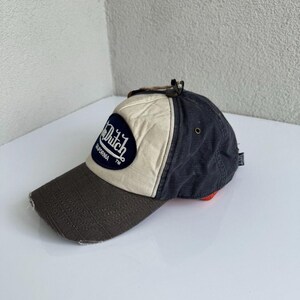 K&ouml;nnte beinhalten: Eine Baseballkappe mit dunkelbraunem Schirm, cremefarbenem Vorderteil und dunkelgrauen Seitenteilen. Ein dunkelblauer, ovaler Aufn&auml;her auf der Vorderseite zeigt den wei&szlig;en Schriftzug Von Dutch California TM. Die Kappe hat eine kleine Metall&ouml;se.
