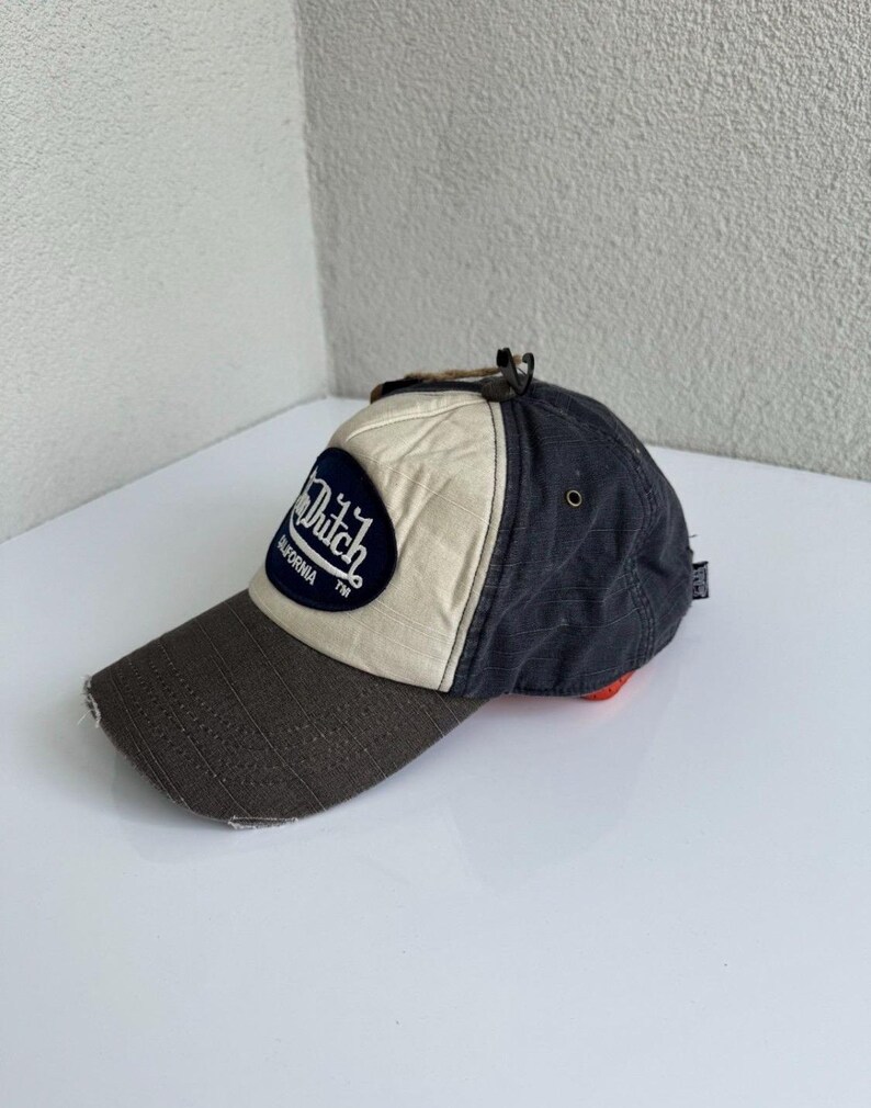 K&ouml;nnte beinhalten: Eine Baseballkappe mit dunkelbraunem Schirm, cremefarbenem Vorderteil und dunkelgrauen Seitenteilen. Ein dunkelblauer, ovaler Aufn&auml;her auf der Vorderseite zeigt den wei&szlig;en Schriftzug Von Dutch California TM. Die Kappe hat eine kleine Metall&ouml;se.
