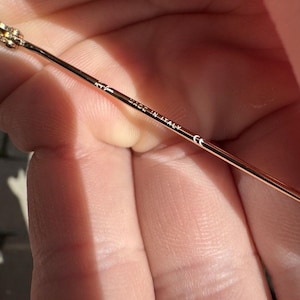 Op de afbeelding: Een dunne, goudkleurige metalen brilpoot. De binnenzijde van de poot is voorzien van het Ray-Ban logo, de tekst "MADE IN ITALY" en het "CE"-keurmerk, gedrukt in kleine, duidelijke witte letters langs het slanke metalen montuur.