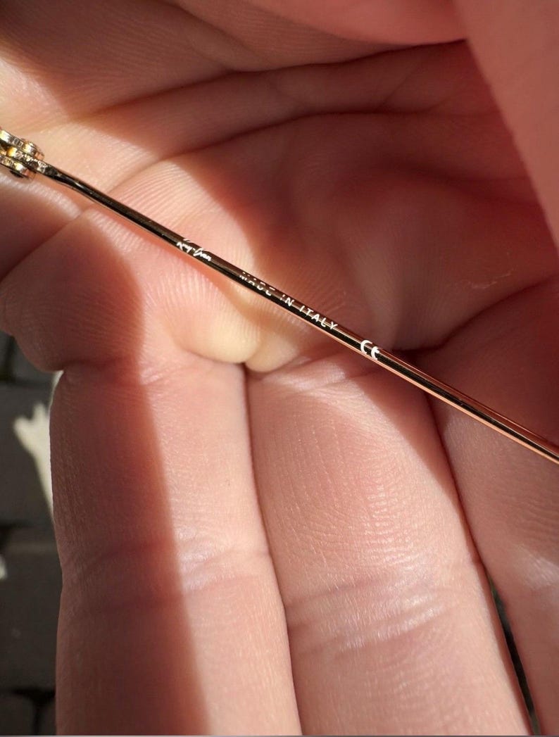 Op de afbeelding: Een dunne, goudkleurige metalen brilpoot. De binnenzijde van de poot is voorzien van het Ray-Ban logo, de tekst "MADE IN ITALY" en het "CE"-keurmerk, gedrukt in kleine, duidelijke witte letters langs het slanke metalen montuur.