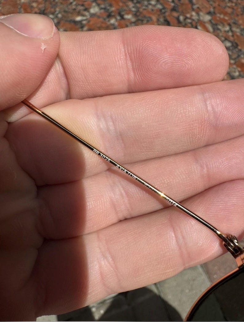 Op de afbeelding: Een dunne goudkleurige metalen brilpoot van een zonnebril. De binnenzijde van de poot is voorzien van een gegraveerde tekst met de tekst RB3025 AVIATOR gevolgd door 58 14. Het metalen oppervlak is glad en reflecterend met een gouden afwerking.