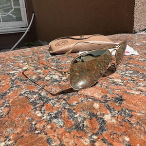 Op de afbeelding: Goudkleurige metalen pilotenzonnebril met reflecterende glazen voorzien van het Ray-Ban logo op het rechterbovenglas. De zonnebril wordt geleverd met een beige beschermetui met ledertextuur, een drukknoopsluiting en een klein wit doekje.