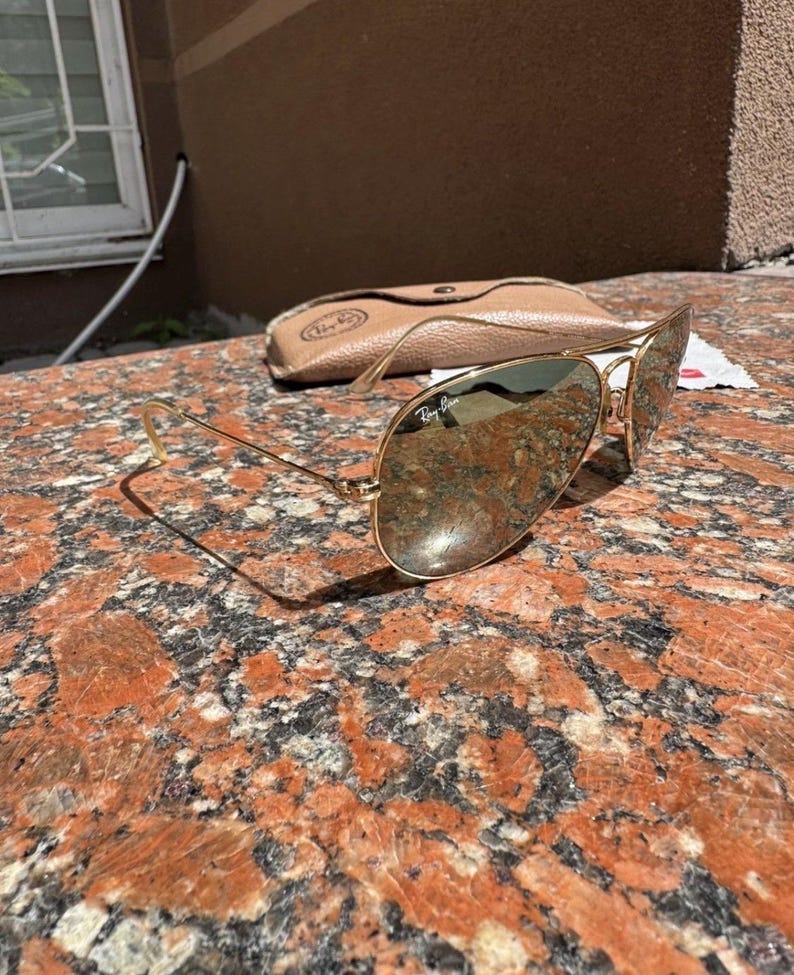 Op de afbeelding: Goudkleurige metalen pilotenzonnebril met reflecterende glazen voorzien van het Ray-Ban logo op het rechterbovenglas. De zonnebril wordt geleverd met een beige beschermetui met ledertextuur, een drukknoopsluiting en een klein wit doekje.