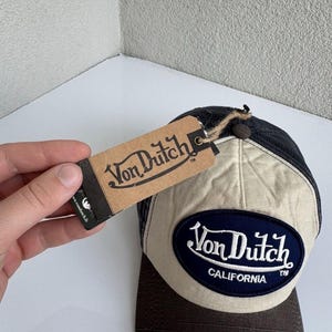 K&ouml;nnte beinhalten: Eine Baseballkappe mit dunkelbraunem Schirm und beigem Vorderteil, das einen dunkelblauen ovalen Aufn&auml;her mit dem wei&szlig;en Schriftzug Von Dutch CALIFORNIA tr&auml;gt. Ein braunes Pappetikett mit dem Von Dutch Logo ist mit einer Kordel befestigt.