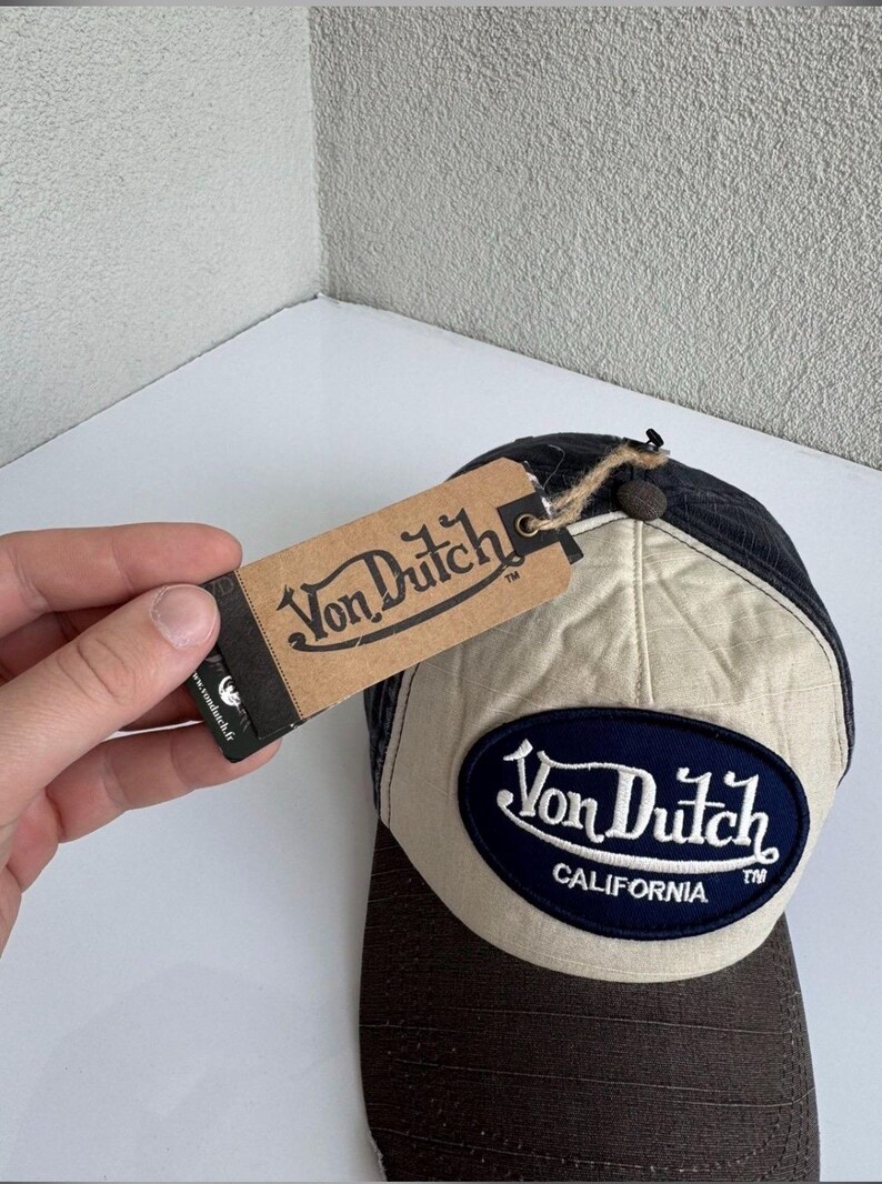 K&ouml;nnte beinhalten: Eine Baseballkappe mit dunkelbraunem Schirm und beigem Vorderteil, das einen dunkelblauen ovalen Aufn&auml;her mit dem wei&szlig;en Schriftzug Von Dutch CALIFORNIA tr&auml;gt. Ein braunes Pappetikett mit dem Von Dutch Logo ist mit einer Kordel befestigt.
