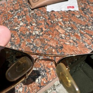Op de afbeelding: Goudkleurige metalen pilotenzonnebril met donkere glazen, verstelbare neuspads en een brug met de inscriptie Ray-Ban 55 14. Inclusief een beige lederen beschermhoes en een wit microvezel reinigingsdoekje met een rood logo.