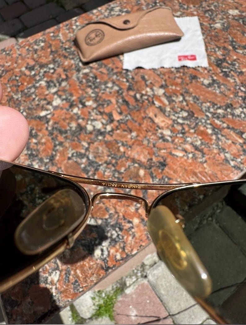 Op de afbeelding: Goudkleurige metalen pilotenzonnebril met donkere glazen, verstelbare neuspads en een brug met de inscriptie Ray-Ban 55 14. Inclusief een beige lederen beschermhoes en een wit microvezel reinigingsdoekje met een rood logo.