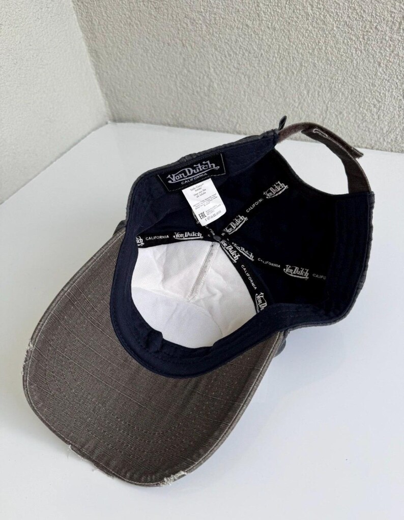 K&ouml;nnte beinhalten: Eine dunkelgraue Baseballkappe mit abgenutztem Schirm, von innen betrachtet. Das Innere verf&uuml;gt &uuml;ber ein schwarzes Schwei&szlig;band mit wei&szlig;em Von Dutch California-Schriftzug, ein wei&szlig;es Stoffetikett und einen braunen verstellbaren Riemen mit Schnalle.